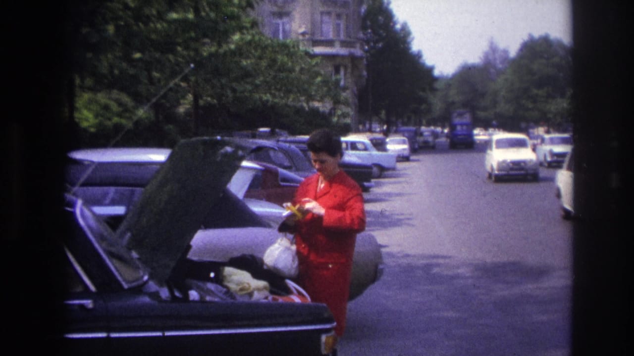 1967 Paris home-movie frame showing a woman in red retrieving items from a parked car trunk on a tree-lined street.