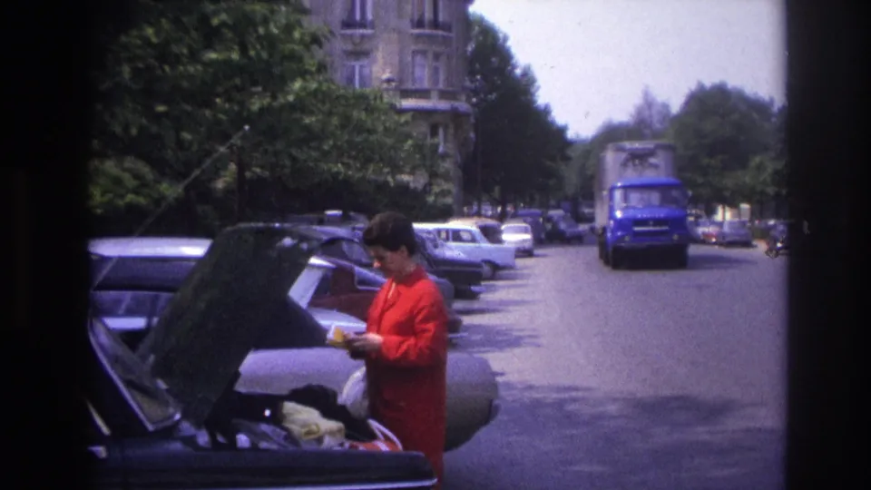 1967 Paris home-movie view of a woman handling belongings at a car trunk with a blue delivery truck approaching behind her.