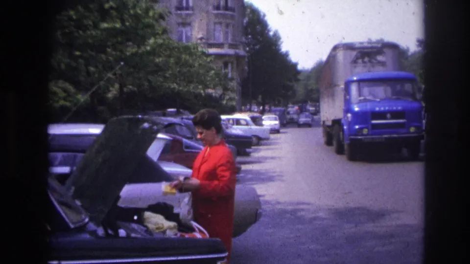 1967 Paris street frame showing a woman in red at a parked car trunk while a blue truck passes close by.
