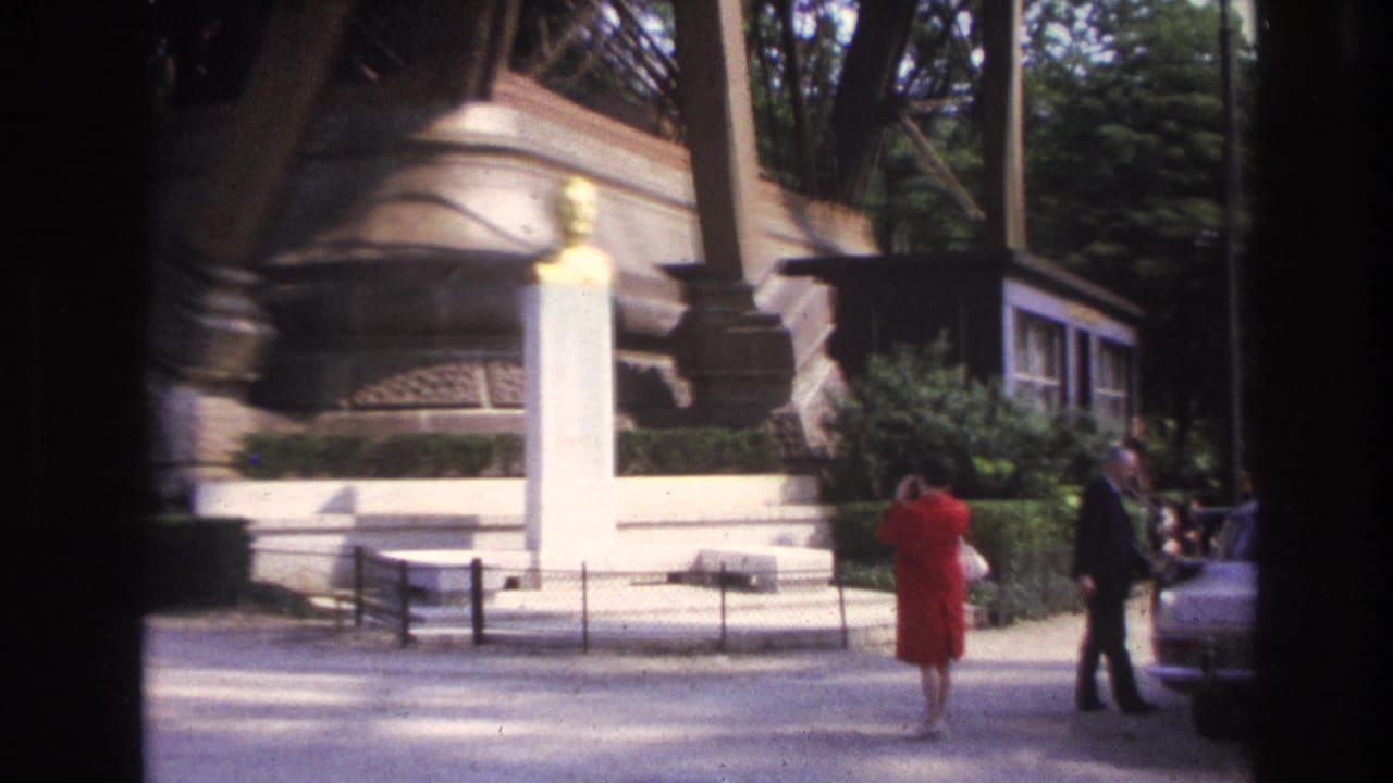 1967 Paris home-movie frame showing a woman in a red dress and a man near a golden bust on a white pedestal by the Eiffel Tower.