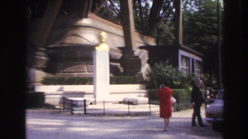 1967 Paris home-movie frame showing a woman in a red dress and a man near a golden bust on a white pedestal by the Eiffel Tower.
