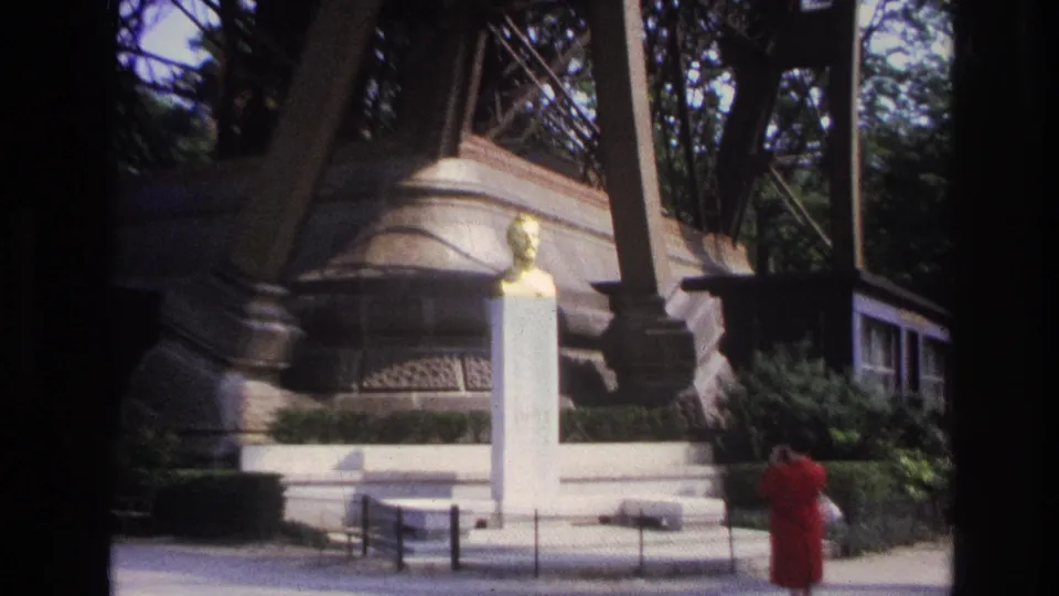 1967 Paris home-movie still centered on a golden bust and white pedestal, with Eiffel Tower ironwork and the woman partly visible nearby.