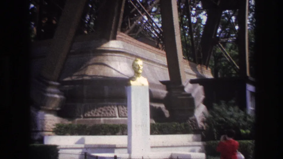 1967 Paris frame showing the golden bust and pedestal in the foreground with the Eiffel Tower structure overhead and a woman in red at right.