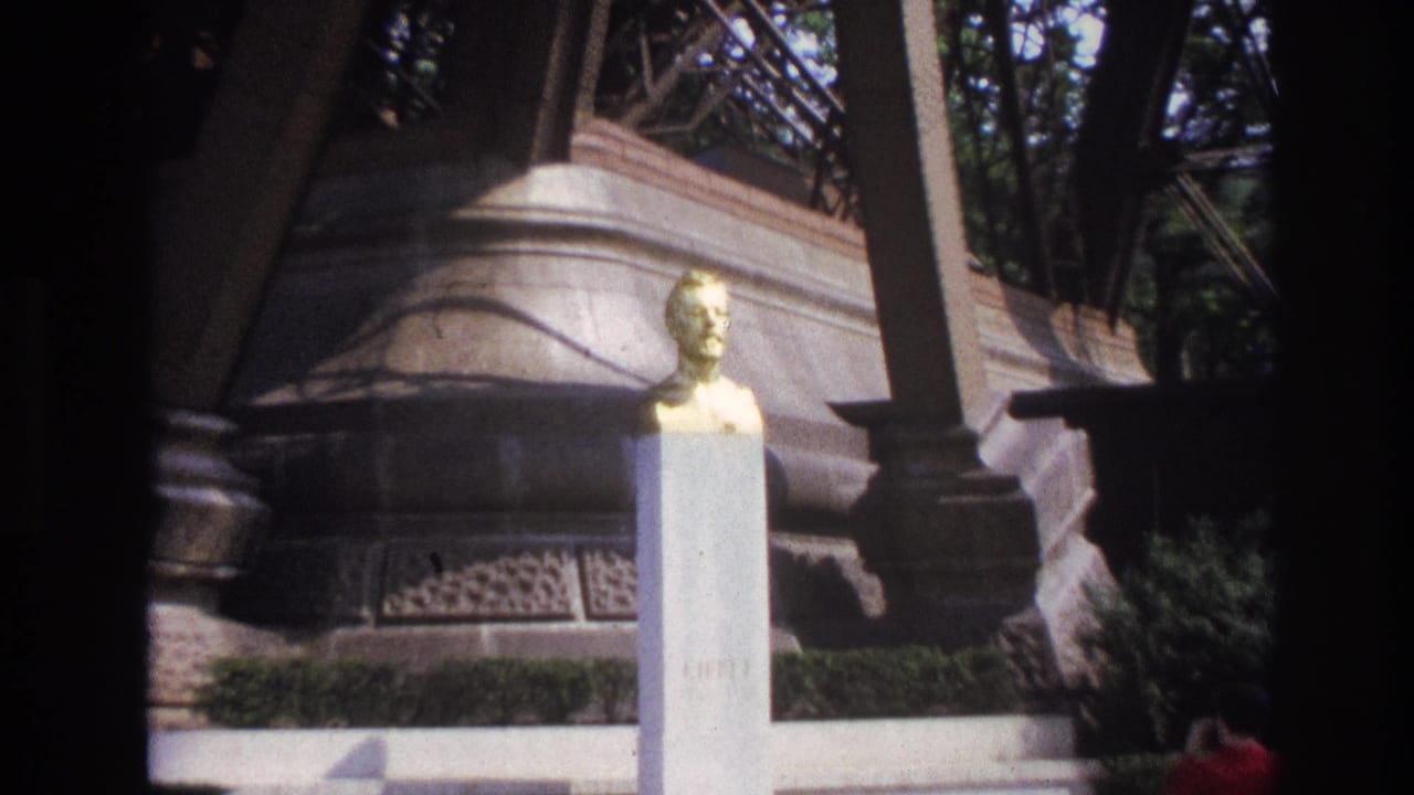 1967 Paris home-movie frame showing a golden male bust on a pale pedestal in front of carved stone architecture and trees.