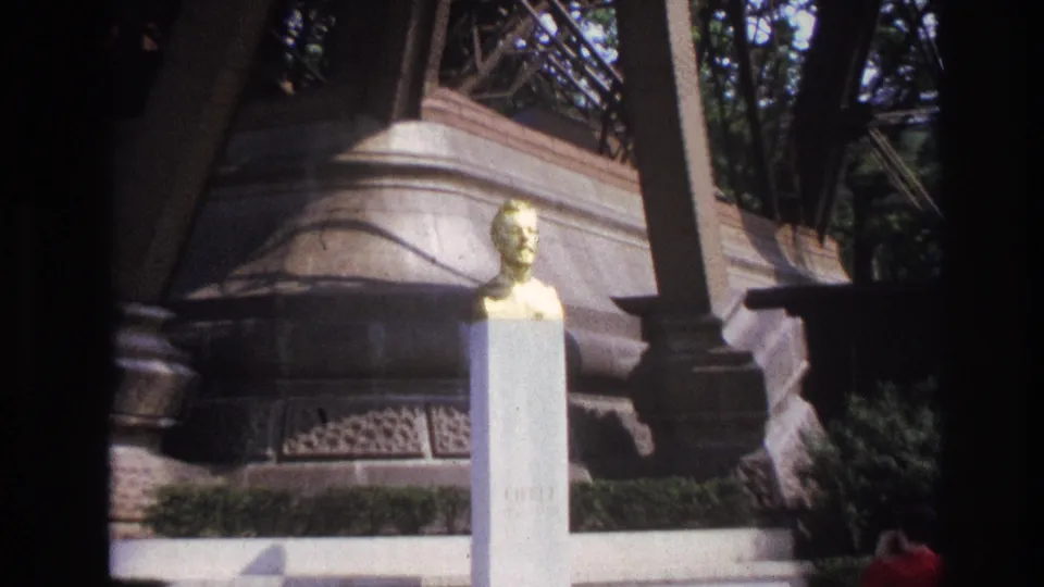 1967 Paris home-movie frame showing a golden male bust on a pale pedestal in front of carved stone architecture and trees.