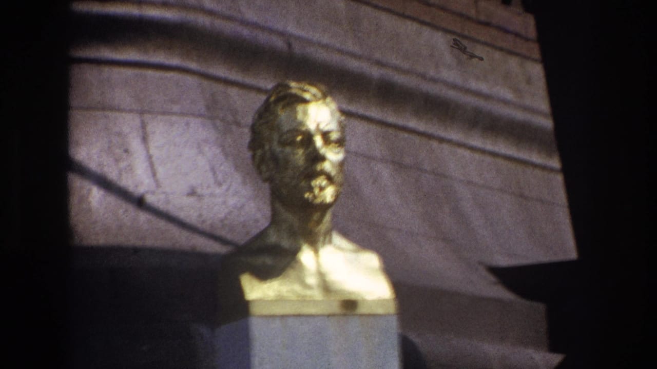 1967 Paris home-movie frame showing a gilded male bust on a stone pedestal with masonry and shadow behind it.