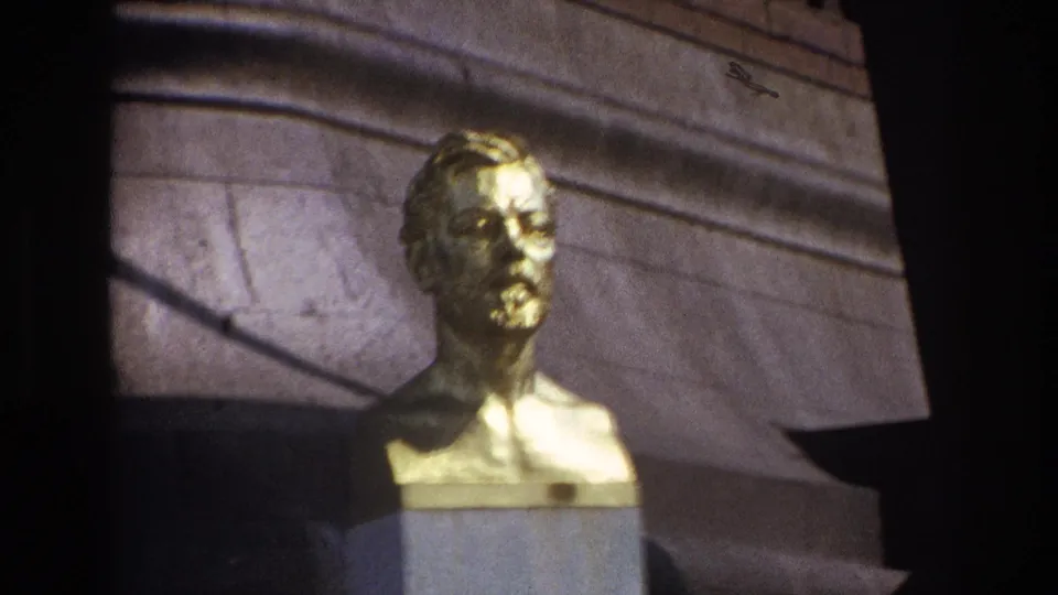 1967 Paris home-movie frame showing a gilded male bust on a stone pedestal with masonry and shadow behind it.