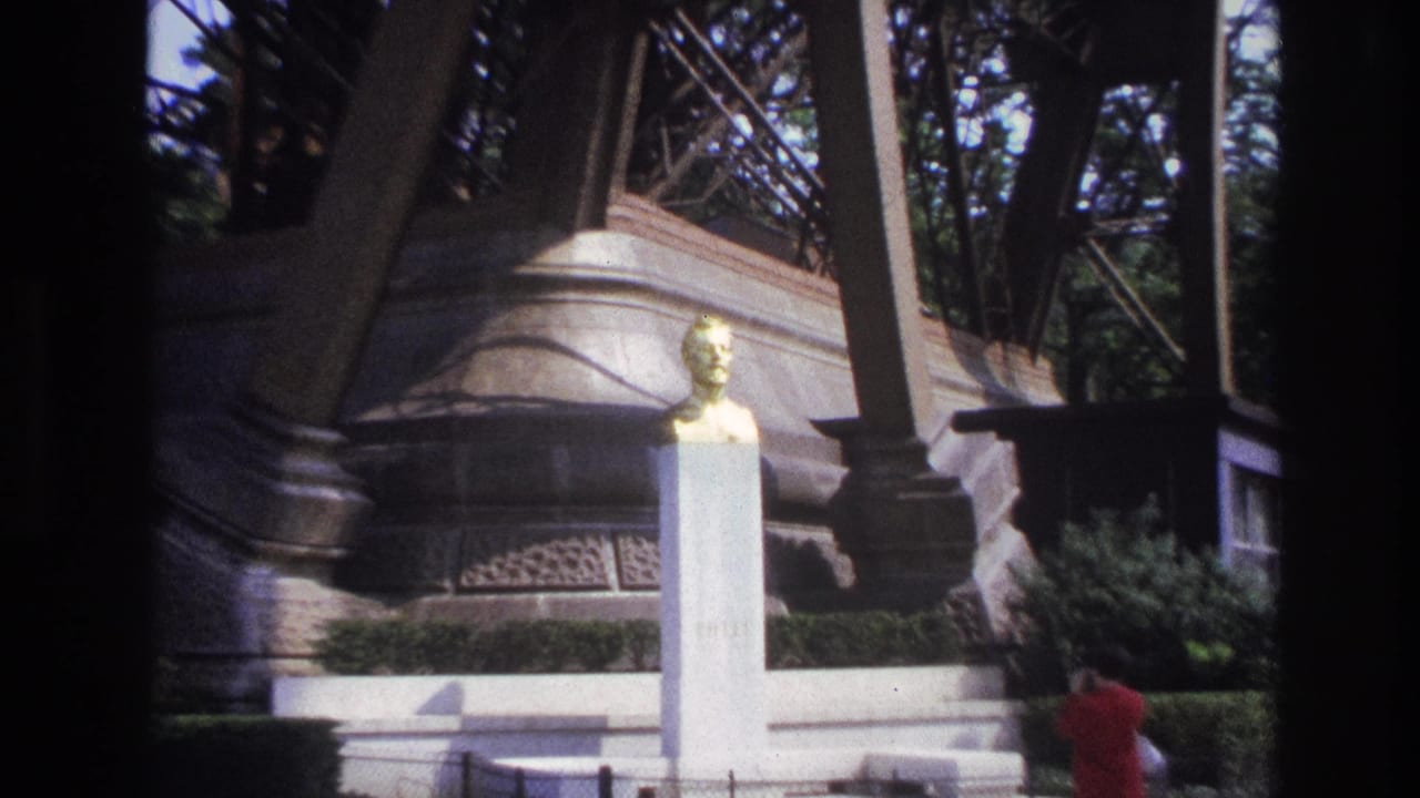 1967 Paris home-movie frame showing a woman in a red dress beside a white monument and golden bust under Eiffel Tower supports.