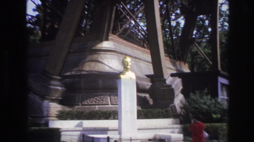 1967 Paris home-movie frame showing a woman in a red dress beside a white monument and golden bust under Eiffel Tower supports.