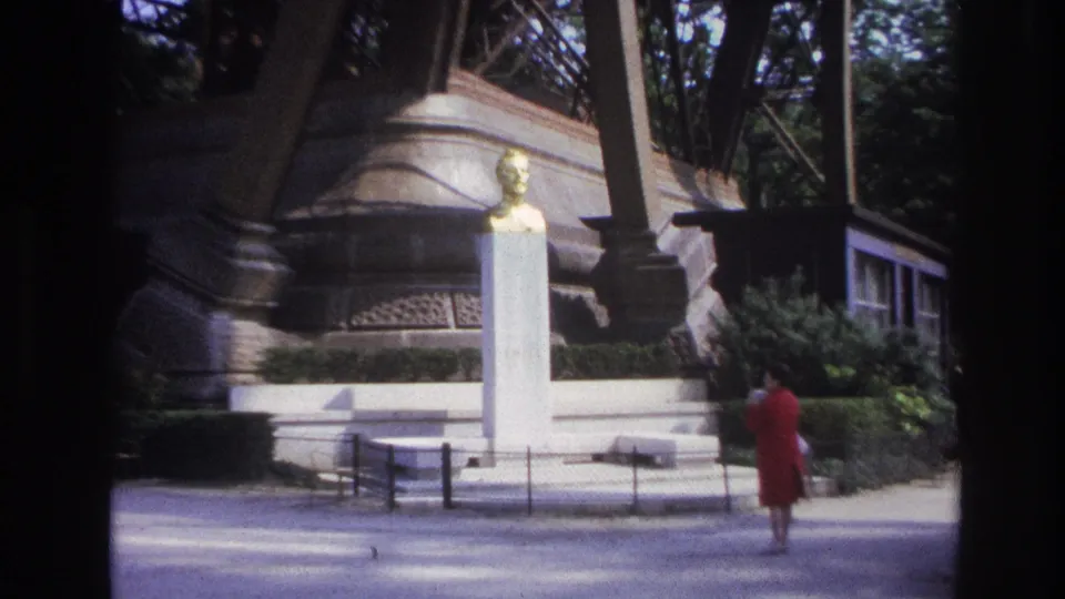 1967 Paris frame with a woman in a red dress walking near a white monument, trees, benches, and iron tower columns.