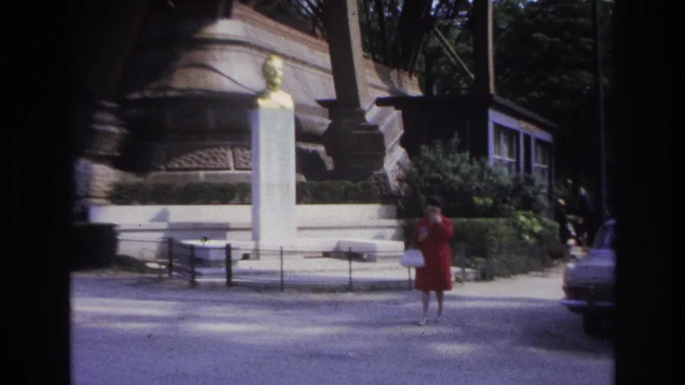 1967 Paris home-movie frame showing a woman in a red dress near a white monument, with a car entering at the right edge.