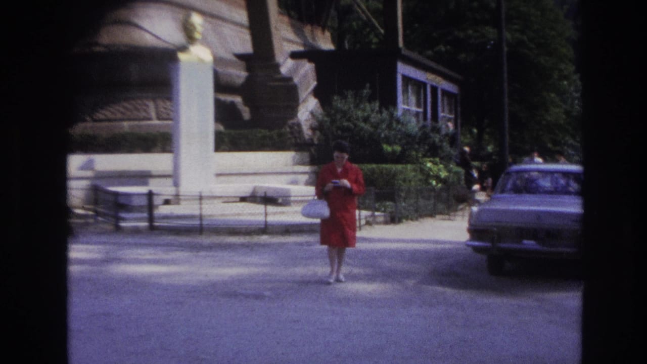 In 1967 Paris footage, a woman in a red dress walks across a street near a stone monument and parked car.