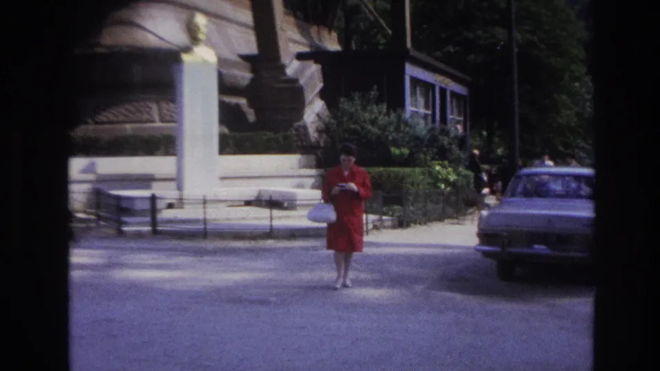 In 1967 Paris footage, a woman in a red dress walks across a street near a stone monument and parked car.
