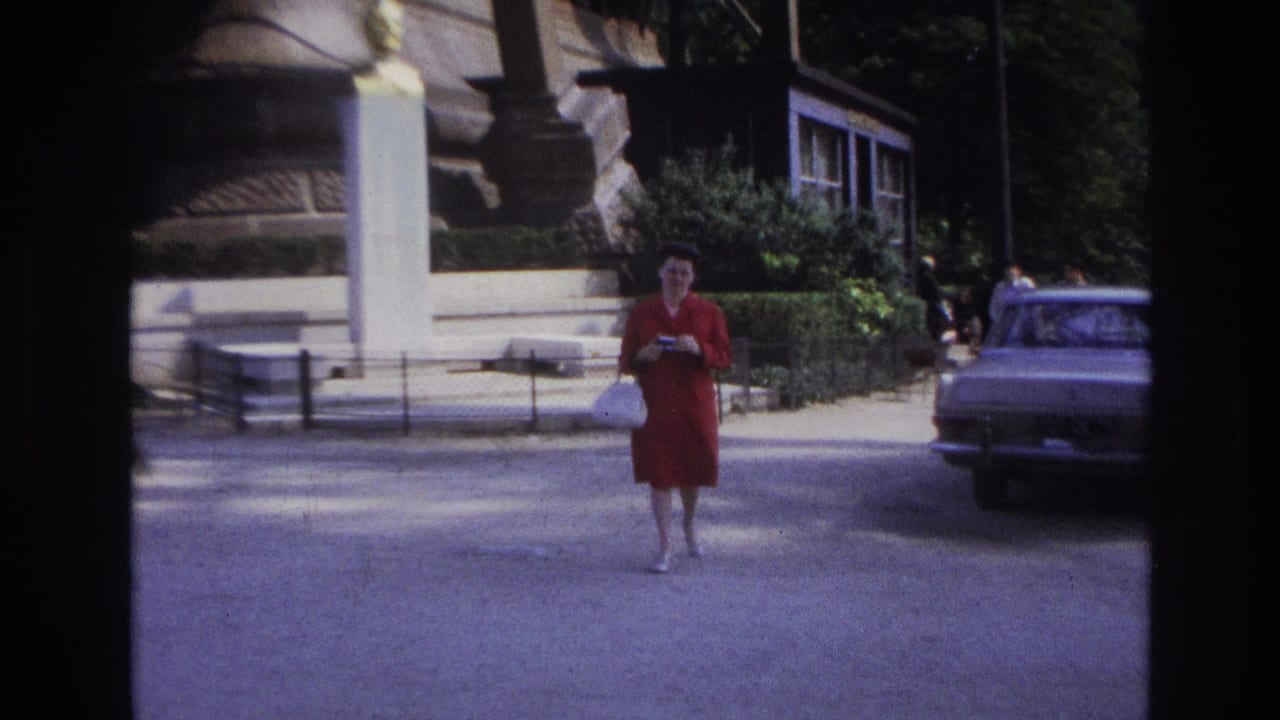 Woman in red continues toward the camera on a street beside a parked car and monument.