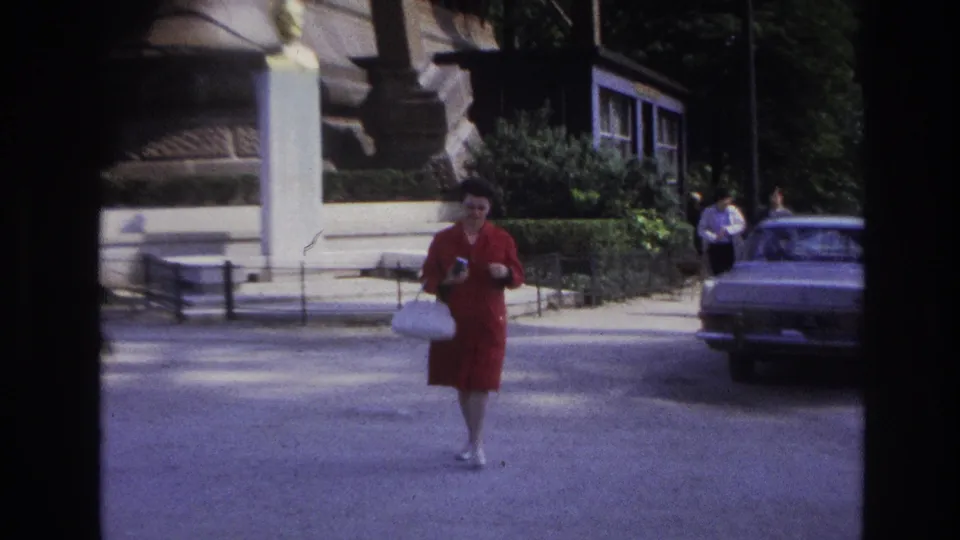 In the 1967 Paris home-movie frame, the woman approaches with her camera as pedestrians appear in the background.