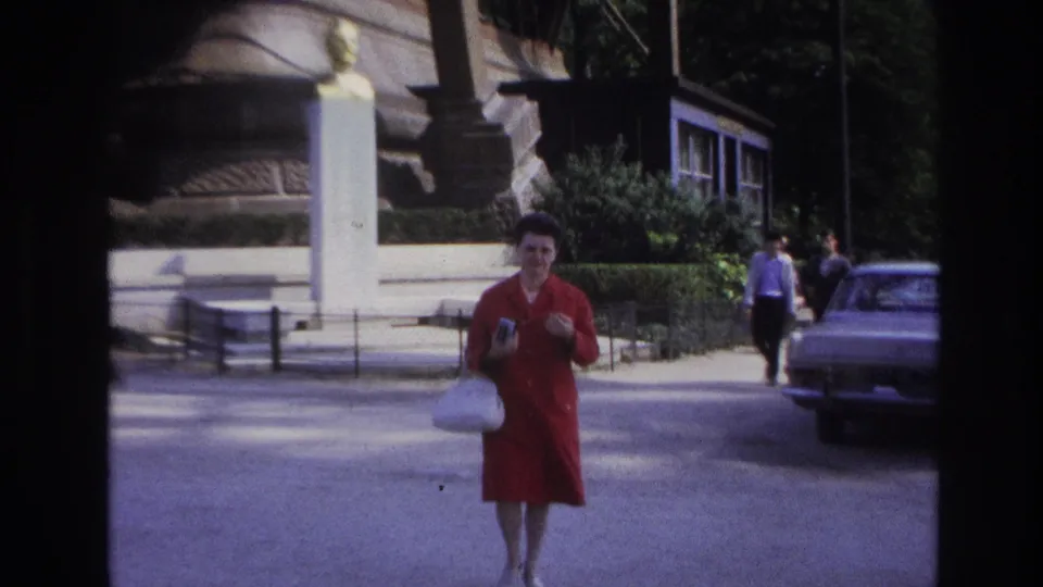 The woman reaches the foreground of the Paris street view, smiling slightly with her camera in hand.