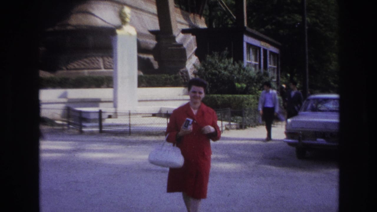 In 1967 Paris home-movie footage, a woman in a red coat walks through a sunlit public area holding a camera and white purse.