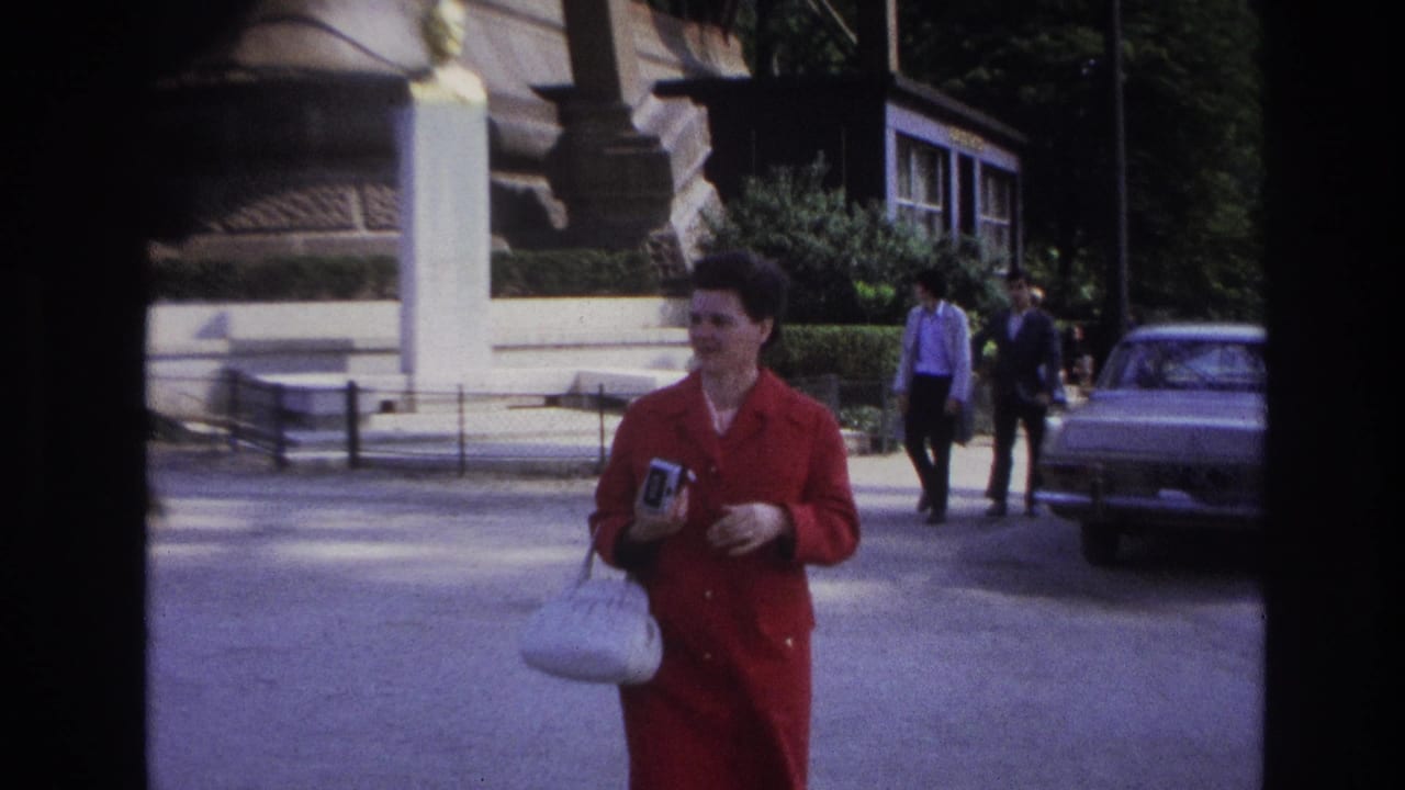 Woman in red coat stands or walks near a monument-like base with pedestrians and a parked car behind her.
