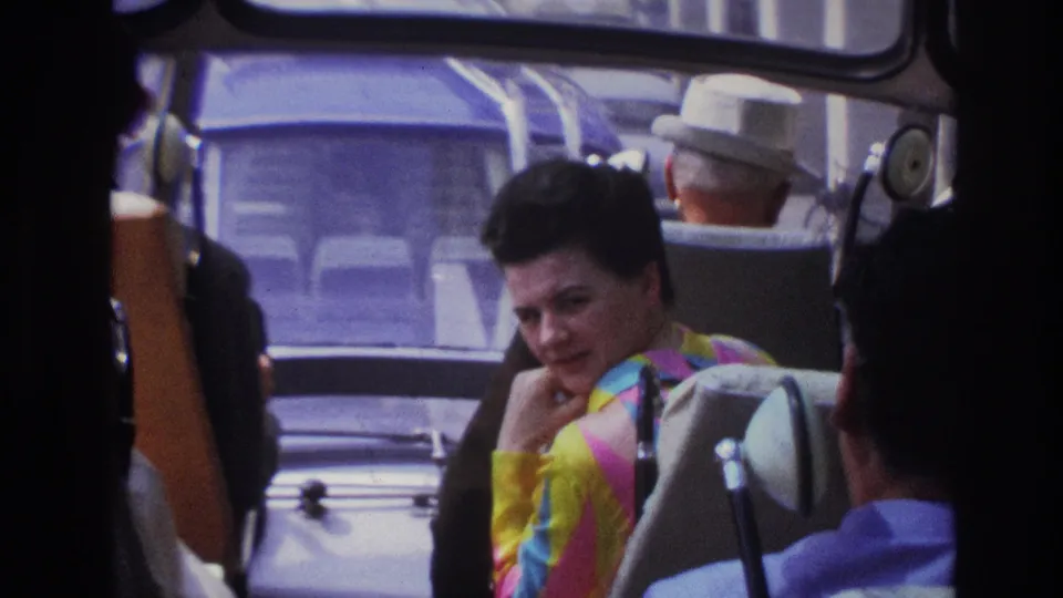 Inside a bus in 1967 Paris footage, the woman wears a colorful striped top and looks back toward the camera from her seat.