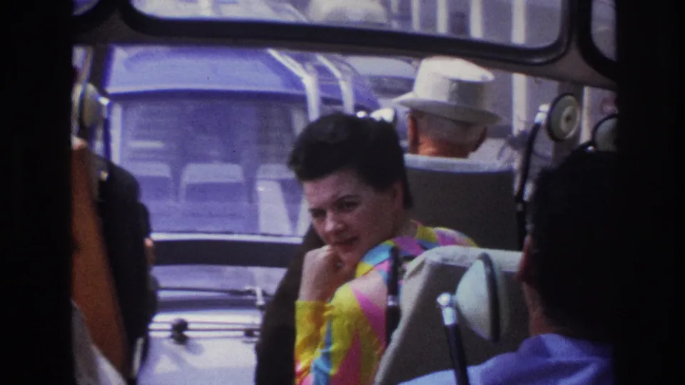 The woman sits inside a Paris bus, framed by seats and windows, with other passengers and a nearby car visible through the glass.