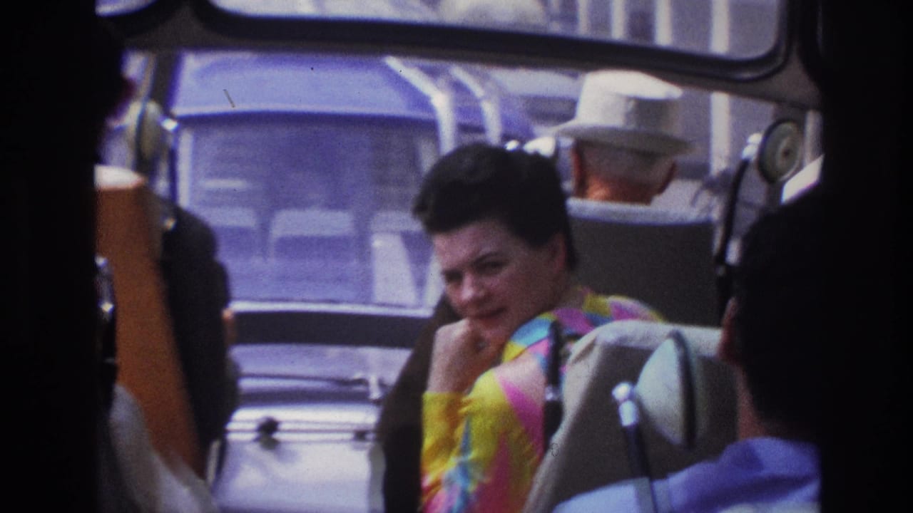 1967 Paris home-movie frame showing a woman in a bright striped top seated on public transport, with passengers and windows around her.