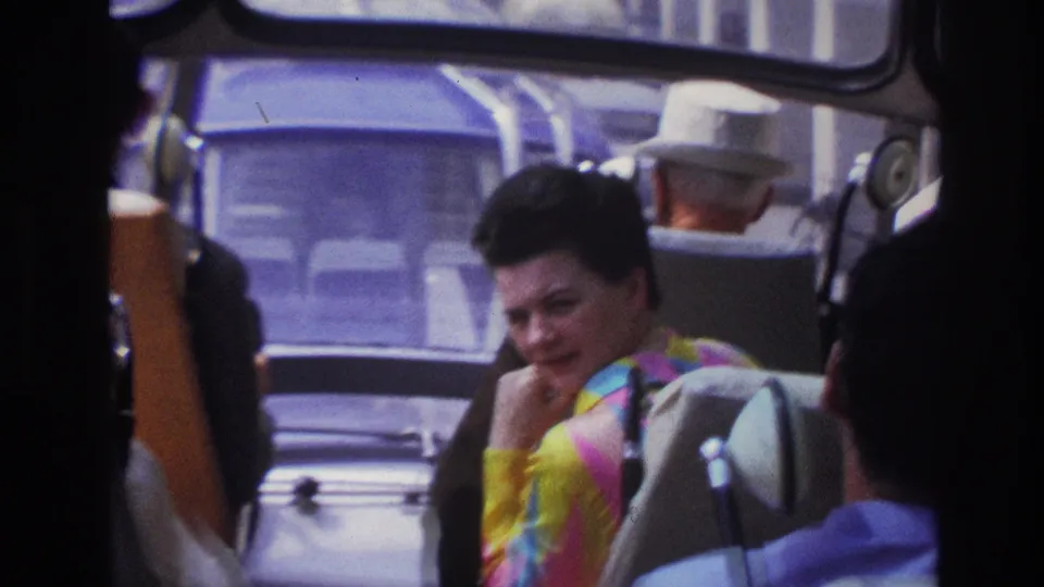 1967 Paris home-movie frame showing a woman in a bright striped top seated on public transport, with passengers and windows around her.