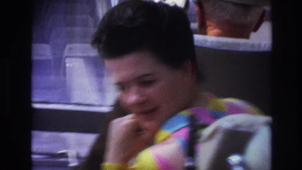 Soft 8mm close view from 1967 Paris showing the woman leaning forward, with the seat back and other passenger shapes partly visible.