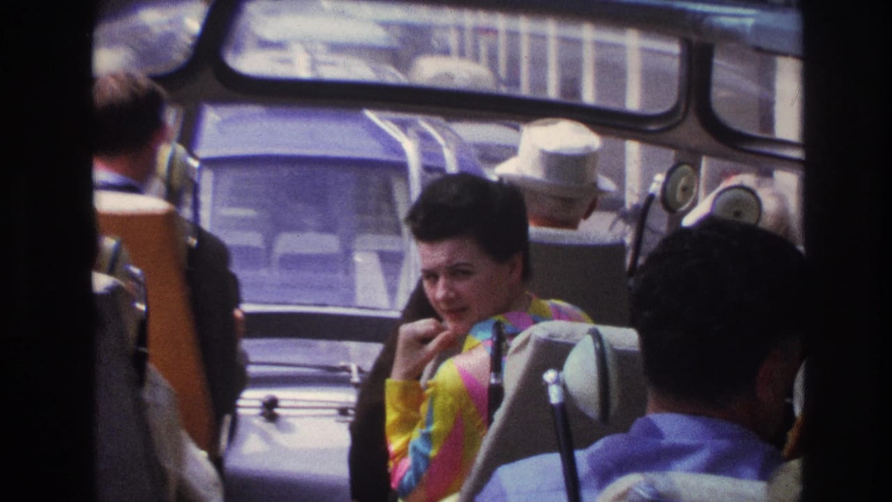 Tour bus passengers seen through seatbacks and windows, with a woman turned toward the camera.