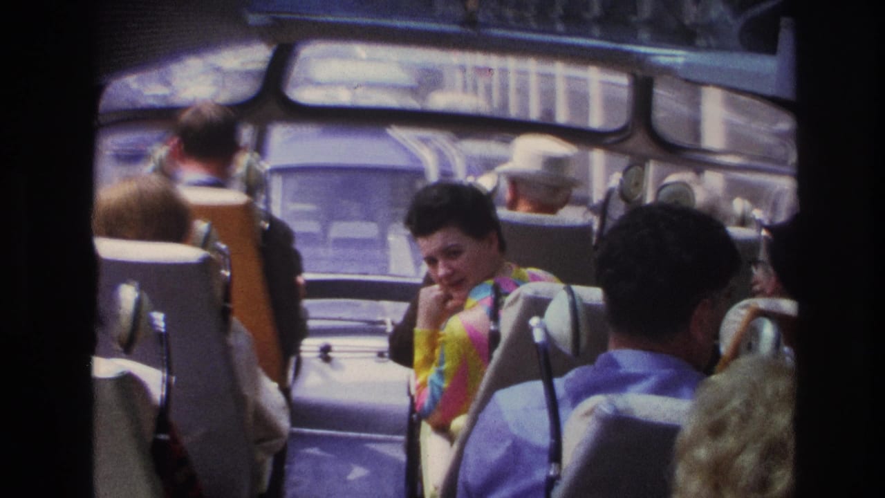 Inside a Paris tour bus in 1967, seated passengers face forward while a woman near the aisle turns toward the camera.
