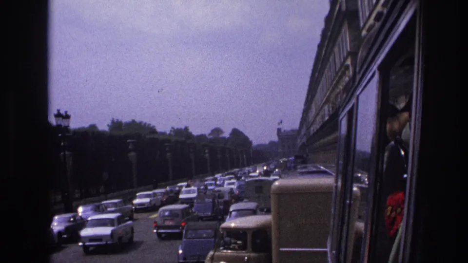 A 1967 Paris roadway crowded with classic cars and vans, filmed from beside or inside a moving tour bus.