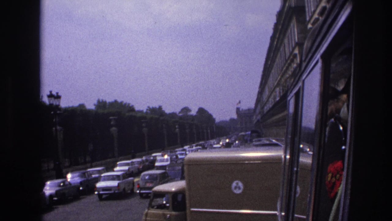 1967 Paris traffic seen from a moving vehicle, with classic cars, a delivery van, roadside lamps, trees, and a bus side at right.