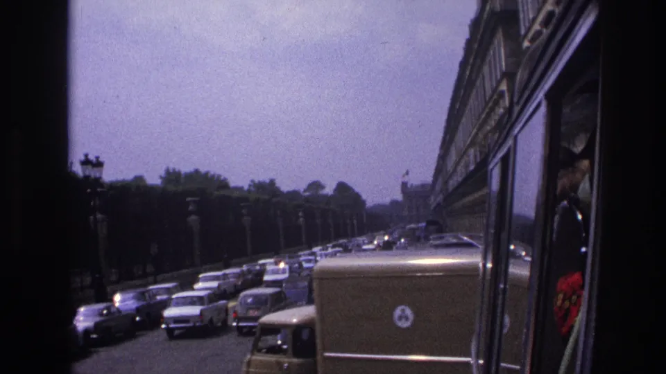 1967 Paris traffic seen from a moving vehicle, with classic cars, a delivery van, roadside lamps, trees, and a bus side at right.