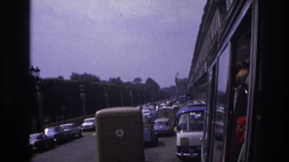 1967 Paris traffic viewed from a moving vehicle, showing a delivery van, classic cars, roadside lamps, trees, and window reflections.