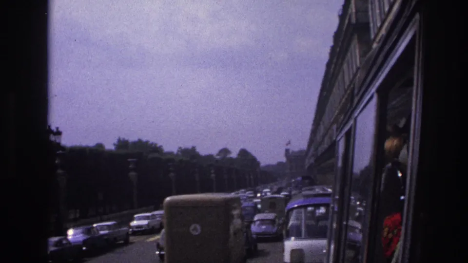 1967 Paris roadway with dense car traffic, a van, a bus front, trees, lamps, and a passenger reflection near the vehicle window.