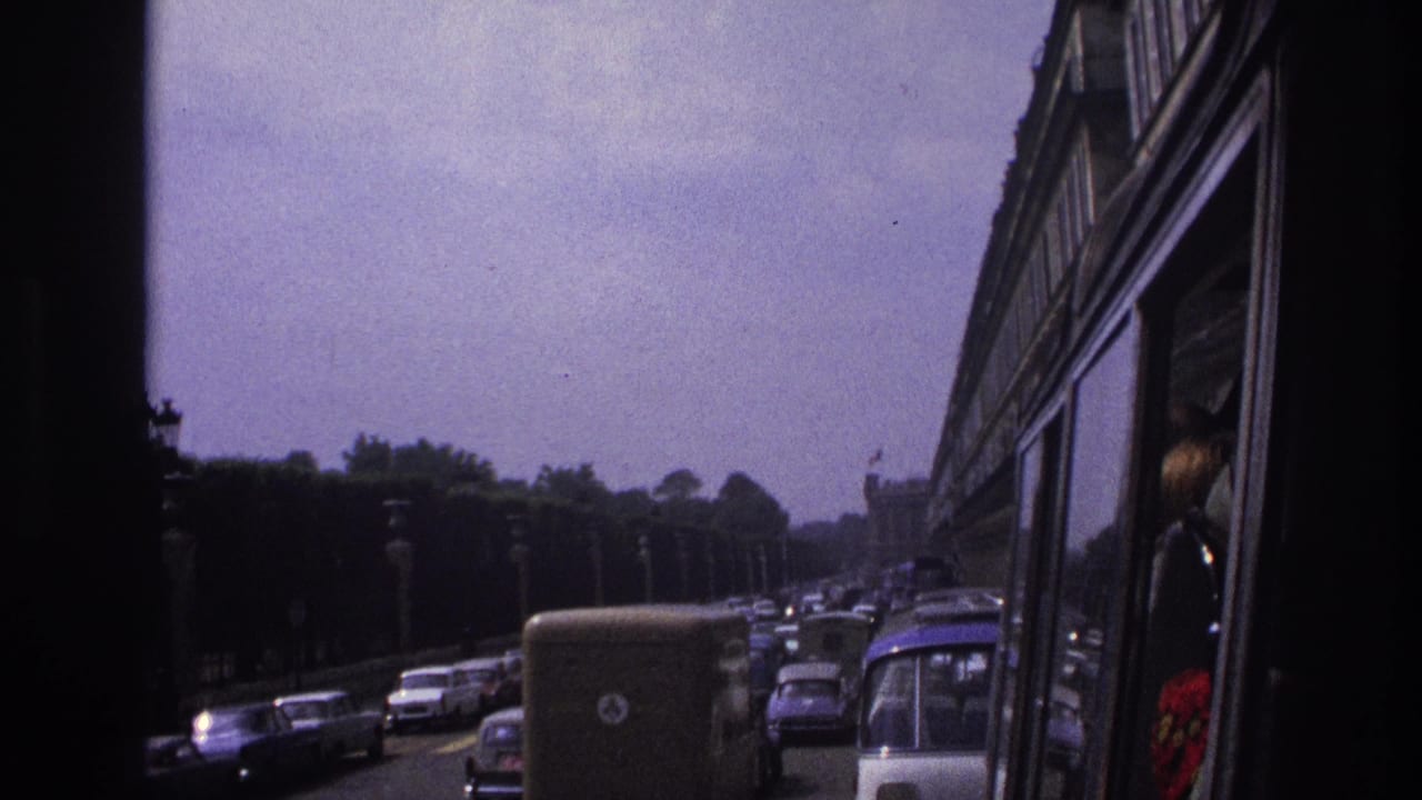 1967 Paris traffic seen from a vehicle, with older cars, a van, roadside lamps, trees, and a bus or coach along the right edge.