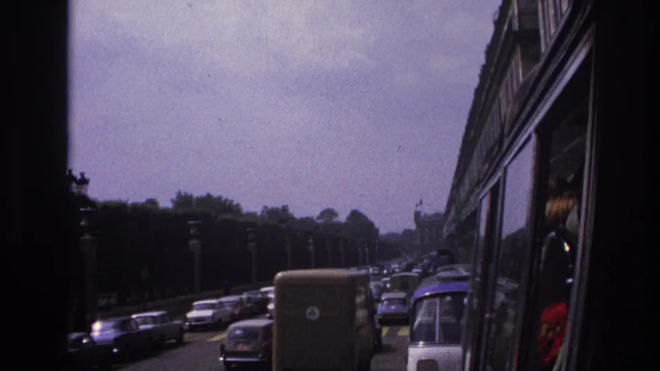 Paris road traffic in 1967, with classic cars, a boxy van, lamps, trees, and a partial passenger visible at the right edge.