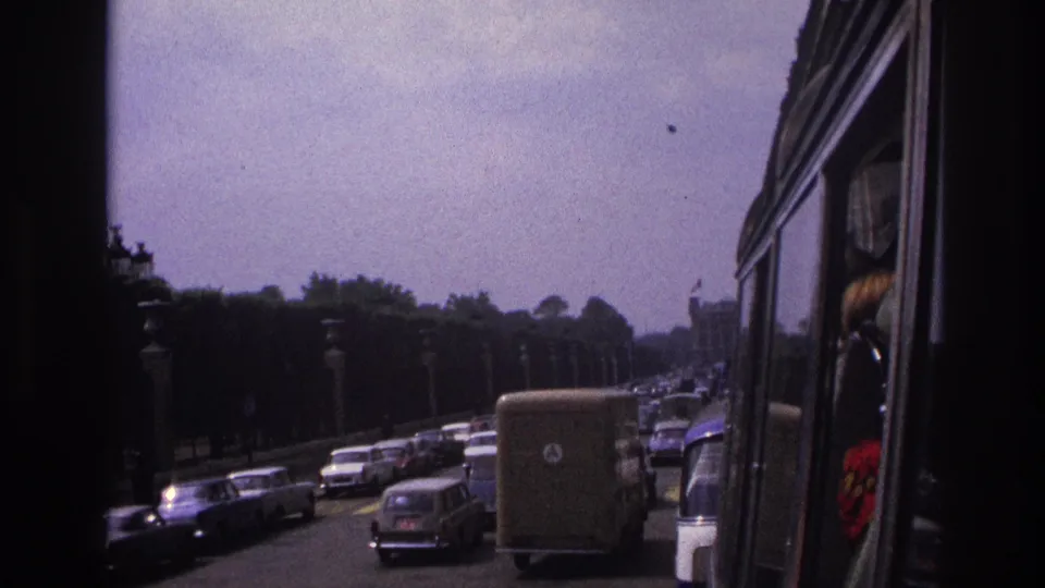 1967 Paris boulevard traffic with older cars, a delivery van, roadside lamps, trees, and the dark edge of the vehicle framing the view.