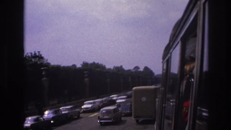 Paris traffic in 1967 seen through a home-movie lens, with older cars, a van, trees, lamps, and part of a bus or coach at frame right.