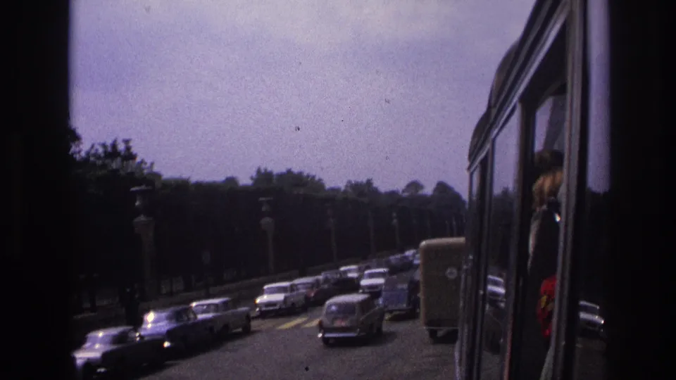 1967 Paris traffic with classic cars on a boulevard, framed by the dark interior edge of a moving vehicle.