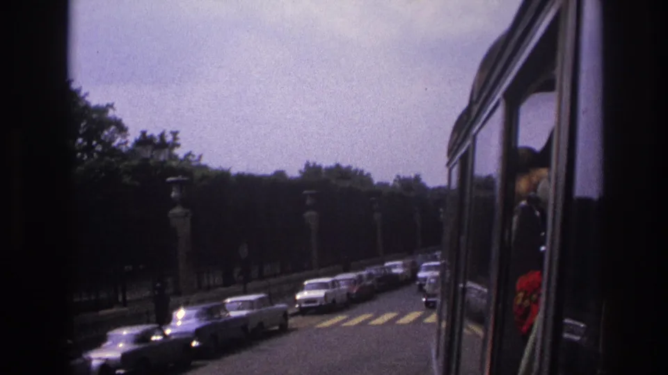 Paris boulevard traffic in 1967, with classic cars receding past trees and lamp posts from a moving-vehicle viewpoint.
