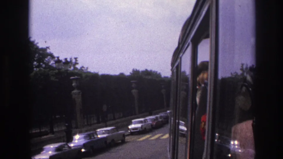 A moving-vehicle view of 1967 Paris traffic, with cars, trees, lamp posts, and a partially visible passenger at the window.