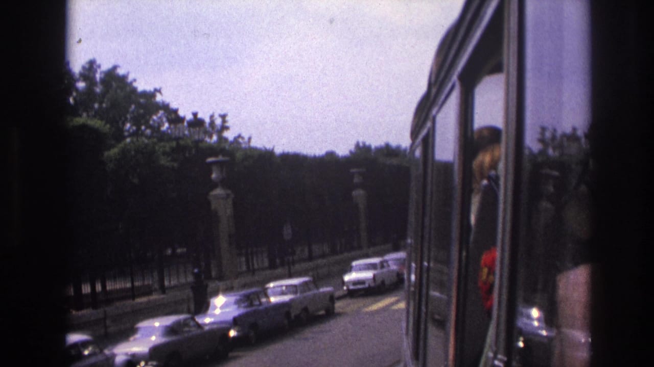 1967 Paris bus-view frame with parked cars, trees, stone pillars, and a passenger reflection at the window edge.