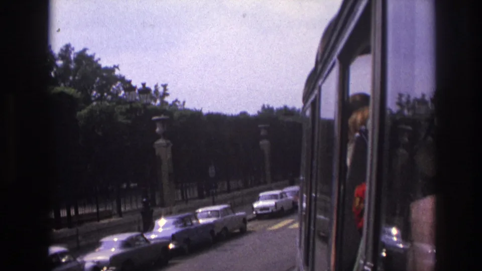1967 Paris bus-view frame with parked cars, trees, stone pillars, and a passenger reflection at the window edge.