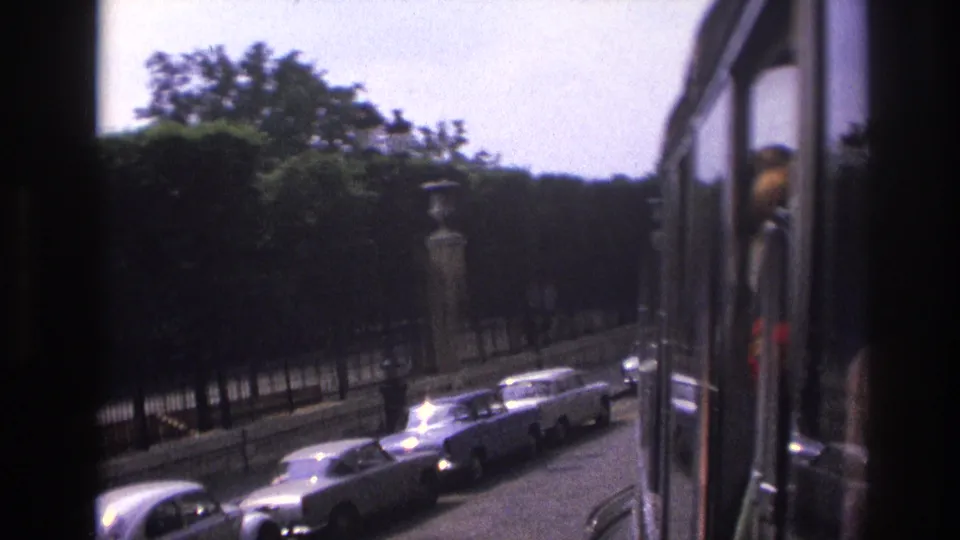 1967 Paris view from the upper deck of a sightseeing bus, showing parked cars, trees, stone pillars, and a reflected passenger.