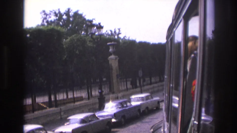 1967 Paris street scene from a sightseeing bus with a row of parked cars, dense trees, stone posts, and window reflections.