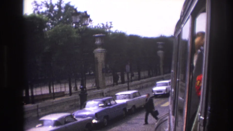 1967 Paris bus-view frame with a suited pedestrian crossing the lower right, parked cars, trees, stone pillars, and a passenger reflection.