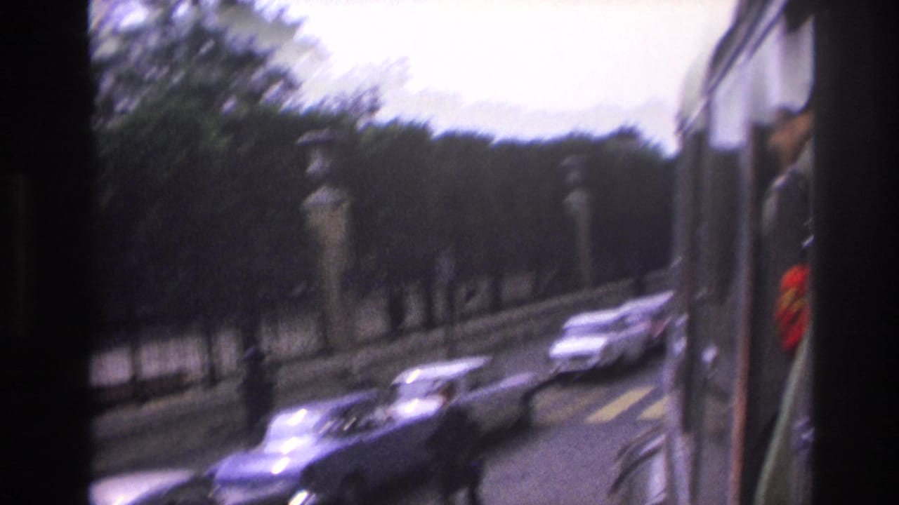 1967 Paris street seen from a moving vehicle, with blurred cars, a pedestrian, stone posts, iron fencing, and roadside trees.