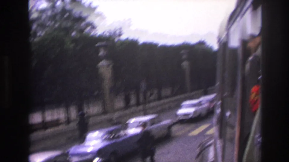 1967 Paris street seen from a moving vehicle, with blurred cars, a pedestrian, stone posts, iron fencing, and roadside trees.