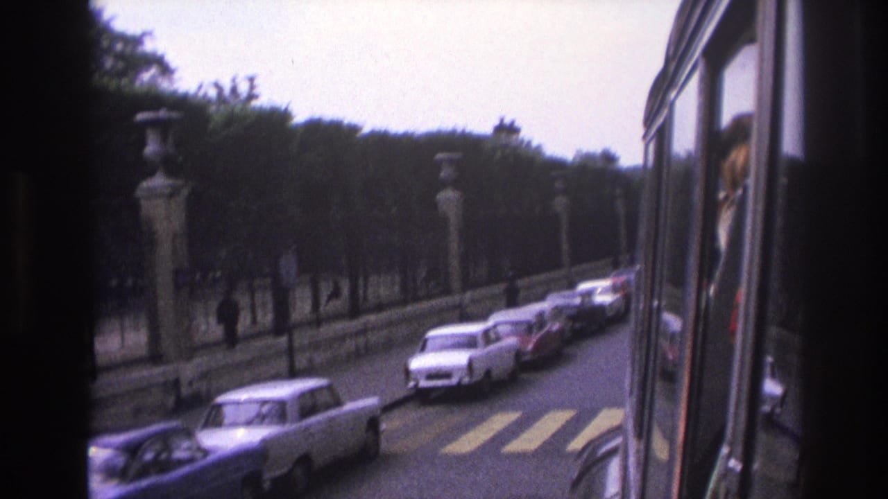 Cars pass along a boulevard beside stone posts, trees, and an iron fence.