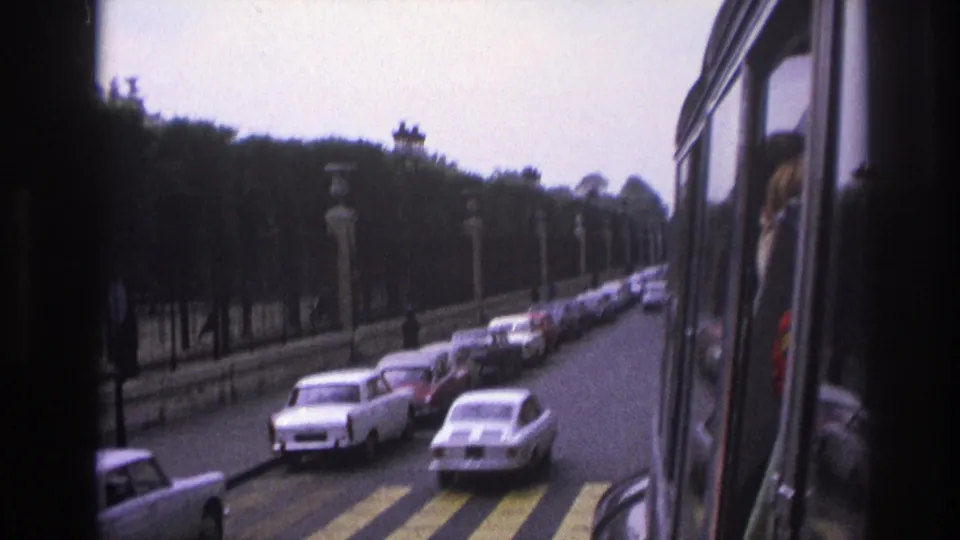 1967 Paris street with parked sedans, a zebra crossing, stone posts, iron fence, trees, and the edge of the filming vehicle.